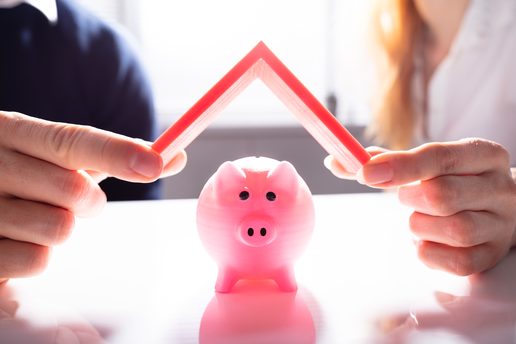 Human Hand Protecting Pink Piggybank With Red Roof Over White Desk